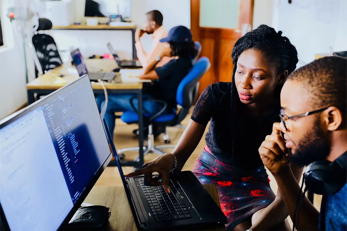 A smiling man works at a computer while his colleague stands watching next to him.