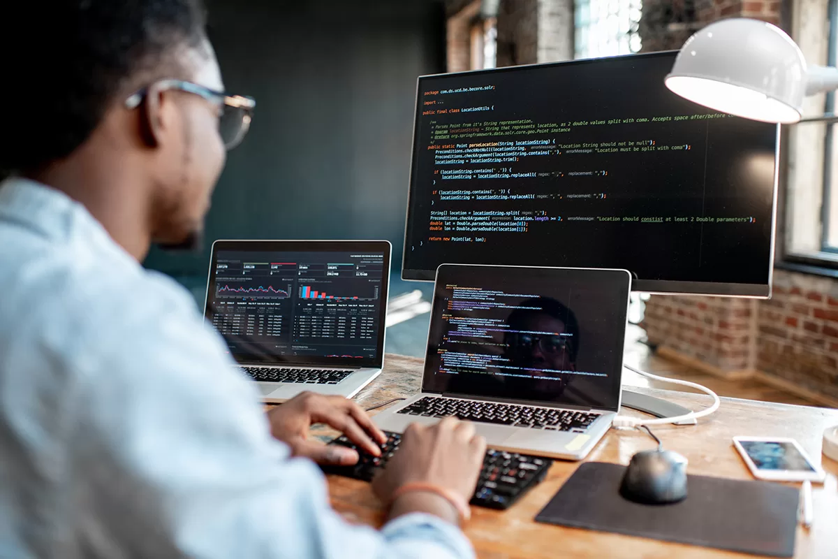 A man wearing glasses looks at three screens on his desk.